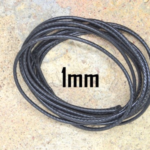 May include: A close-up of a black, braided cord, approximately 1 mm thick, coiled on a light brown surface. The cord is made of a flexible material and is suitable for crafting projects.