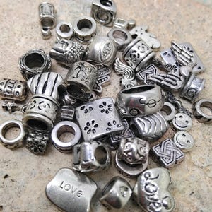 Op de afbeelding: Een verzameling diverse zilverkleurige metalen kralen met verschillende vormen en ontwerpen, waaronder een vierkante kraal met bloemmotieven, een hartvormige kraal met het woord LOVE, een smiley-kraal en diverse cilindrische tussenkralen.