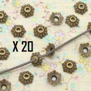 May include: Twenty antique gold-toned metal flower-shaped bead caps. The bead caps have a raised design and a hole in the center. The bead caps are arranged on a fabric surface with a brown cord running through some of them. The text "X 20" is printed on the fabric.