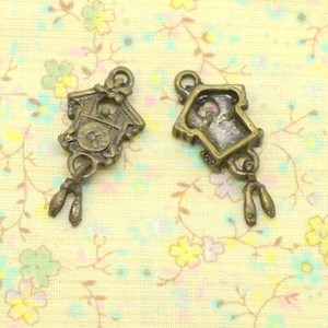 May include: Two bronze-colored clock charms. Each charm is a miniature cuckoo clock with a detailed face and pendulum. The charms are designed for jewelry making or crafting projects. The background is a light yellow fabric with a floral pattern.