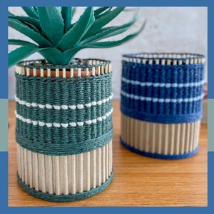 May include: Two decorative woven planters. One is teal with white accents and a natural wood base, holding a green plant. The other is blue with white accents and a natural wood base. Both are cylindrical and sit on a wooden surface.