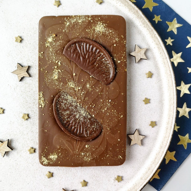 Puede incluir: Una barra de chocolate con dos segmentos de naranja en la parte superior. El chocolate est&aacute; cubierto de chispas doradas.