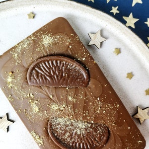 Puede incluir: Primer plano de una barra de chocolate con un dise&ntilde;o decorativo de rodajas de naranja. El chocolate est&aacute; cubierto con una capa de chispas doradas.