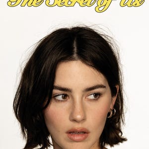 May include: A portrait featuring the text The Secret of Us in yellow cursive font above a person with dark brown hair, brown eyes, and a small gold hoop earring in the left ear. The person has a neutral expression and is looking to the right.