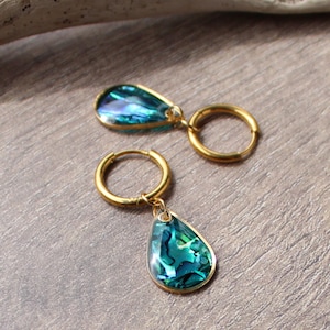 Peut inclure: Une paire de boucles d'oreilles créoles dorées avec des pendentifs en forme de goutte. Les pendentifs présentent des coquillages d'ormeau bleu-vert irisés, encadrés d'une bordure dorée. Les boucles d'oreilles sont présentées sur une surface texturée de ton neutre.