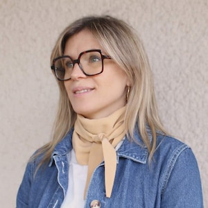 Peut inclure: Une femme portant de grandes lunettes carrées en écaille de tortue et des créoles dorées. Elle porte une écharpe beige clair nouée au cou et une veste en jean. Ses cheveux blonds sont séparés sur le côté.