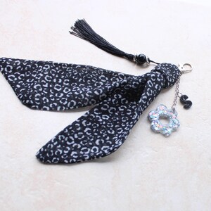 Peut inclure: Un accessoire en tissu à motifs noir et blanc avec un pompon et des breloques décoratives. L'accessoire comprend un fermoir argenté, un charm en forme de fleur avec des paillettes irisées et un charm noir en forme de lettre "S".