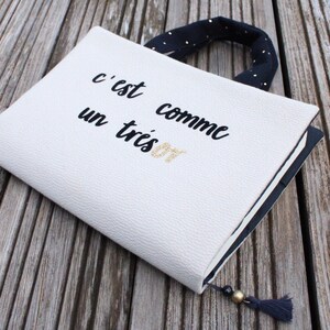 May include: A white notebook cover with black text that reads "c'est comme un tr&egrave;s bon". The cover has a black and white polka dot handle.