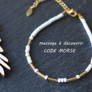 Peut inclure: Un bracelet délicat composé de perles de rocaille blanches et de perles cylindriques dorées. Le bracelet comprend une chaîne et un fermoir dorés. Le texte "message à découvrir CODE MORSE" est affiché en blanc.