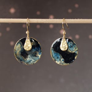 Peut inclure: Une paire de boucles d'oreilles pendantes dorées. Chaque boucle d'oreille présente un disque circulaire bleu foncé et doré, avec un charm en forme de goutte d'eau doré texturé. Les boucles sont suspendues à des crochets dorés.