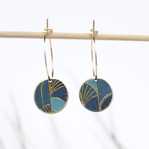 Peut inclure: Boucles d'oreilles créoles dorées avec un pendentif rond bleu et or. Le motif présente des formes géométriques et des lignes.