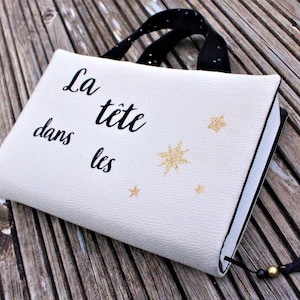 May include: A white book cover with a black strap and a gold tassel. The cover has the text "La t&ecirc;te dans les" and gold glitter stars.