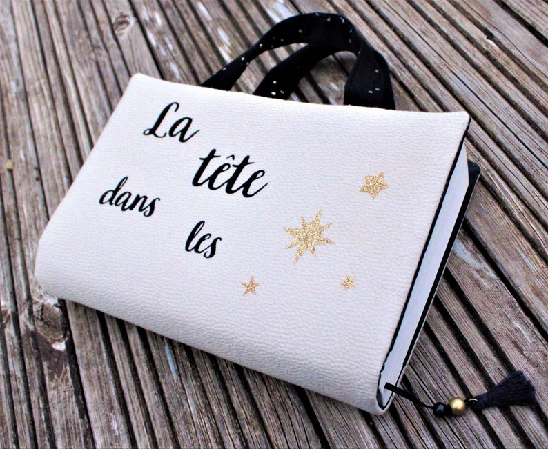 May include: A white book cover with a black strap and a gold tassel. The cover has the text "La t&ecirc;te dans les" and gold glitter stars.