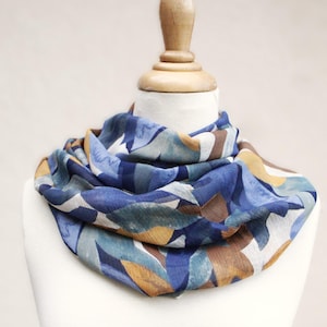 Peut inclure: Écharpe infinie à motifs bleus, marron et jaunes, présentée sur un mannequin blanc. Le motif de l'écharpe est de type aquarelle avec des formes de feuilles bleues, marron et jaunes. L'écharpe est faite d'un matériau léger.