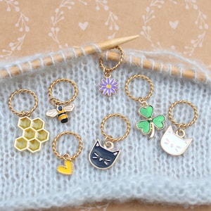 Peut inclure: Ensemble de marqueurs de mailles dorés avec différents breloques : nid d'abeille, abeille, cœur, chat, trèfle et fleur. Les breloques sont jaunes, noires, blanches, vertes et violettes. Sur un tissu tricoté bleu.
