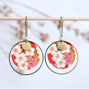 Peut inclure: Une paire de boucles d'oreilles créoles dorées avec un motif floral rouge et blanc et un charme rond doré. Les boucles d'oreilles sont suspendues à une baguette en bois.