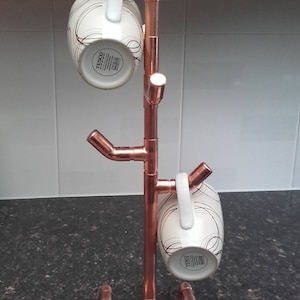 May include: Copper mug rack with two white mugs hanging on it. The rack is made of copper pipe and has a tree-like design. The mugs have black and white designs on them.