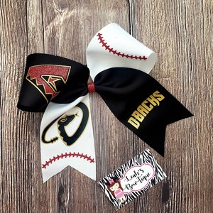 May include: A black and white cheer bow with a red and gold Arizona Diamondbacks logo on one side and a black and gold snake logo on the other side. The bow has a white baseball stitch design.