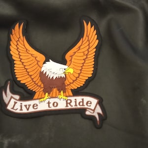 Peut inclure: Une veste en cuir noire avec un écusson brodé représentant un aigle aux ailes déployées et une bannière sur laquelle est inscrit "Live to Ride".