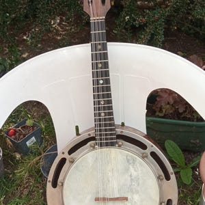 Peut inclure: Un banjo-mandoline vintage avec un manche et une tête en bois, et un corps rond cerclé de métal. L'instrument a des cordes et est posé sur une chaise blanche, avec de la verdure en arrière-plan.