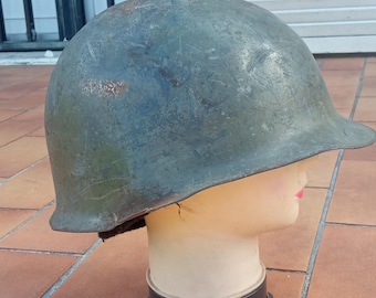 May include: An olive drab military helmet, likely from the mid-20th century, is displayed on a mannequin head. The helmet shows signs of wear, with a slightly weathered appearance. The mannequin head has a neutral skin tone.