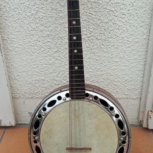 Peut inclure: Un banjo-mandoline vintage avec un manche et une tête en bois, et un corps rond avec une jante en métal. L'instrument a des cordes et des chevilles d'accord, et est posé contre un mur blanc texturé et un sol en carrelage de terre cuite.