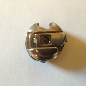 Puede incluir: Una canillera metálica brillante para máquina de coser. La canillera tiene forma redondeada con una abertura rectangular y un pequeño orificio circular. El metal tiene una superficie reflectante.