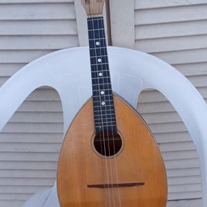 Peut inclure: Mandoline vintage en bois brun clair, posée sur une chaise blanche. L'instrument a une rosace ronde et une touche avec des incrustations de points blancs. La tête comporte des chevilles d'accord.