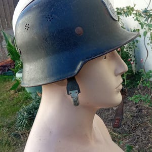 May include: A black metal helmet with a silver-coloured top section, featuring a series of small ventilation holes along the side, a metallic emblem on the front, and a black chin strap with a silver metal buckle, displayed on a mannequin head.