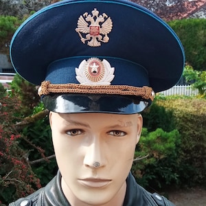 May include: A dark blue military-style peaked cap featuring a gold-toned double-headed eagle emblem on the crown and a silver-toned star badge on the front band. The cap includes a black visor and a decorative gold braided cord across the front.