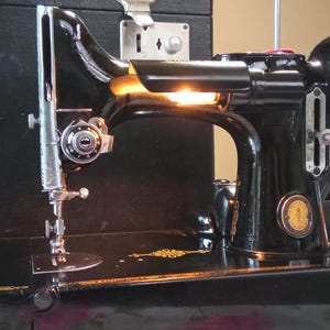 Pode incluir: Máquina de costura Singer preta vintage com agulha e detalhes prateados. A máquina está sobre uma base preta, com uma caixa preta à esquerda. A máquina tem um emblema dourado e é iluminada por dentro.
