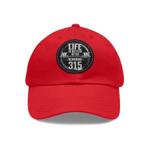 May include: A bright red baseball cap with a curved brim. The cap features a circular patch with the text "LIFE IS BETTER AFTER BENCHING 315" around a barbell graphic. The text and graphic are in white and gray.
