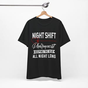 Può includere: Una t-shirt nera a maniche corte con testo bianco che recita NIGHT SHIFT PHARMACIST KEEPING THE BEAT ALL NIGHT LONG. Un grafico a cuore rosso con una linea EKG che lo attraversa è posizionato sopra la parola Pharmacist sul davanti.