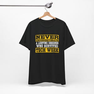 May include: Black t-shirt with the text "NEVER UNDERESTIMATE A LIGHTING DESIGNER WHO SURVIVED TECH WEEK" in yellow and white block letters. The shirt is hanging on a wooden hanger.