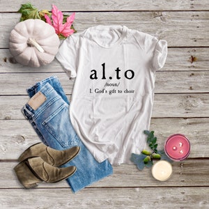 Könnte beinhalten: Weißes T-Shirt mit dem Text "al.to /Substantiv/ 1. Gottes Geschenk an den Chor" darauf gedruckt. Das Shirt liegt auf einer Holzoberfläche mit einer blauen Jeans, zwei braunen Stiefeln, einem weißen Kürbis und zwei Kerzen.