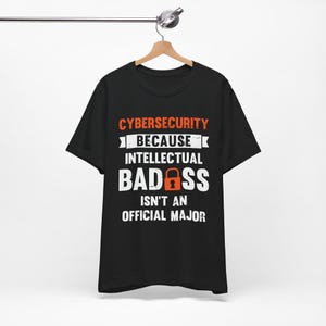 May include: Black t-shirt with the text "CYBERSECURITY BECAUSE INTELLECTUAL BADASS ISN'T AN OFFICIAL MAJOR" in white and orange lettering. The word "BADASS" features a red padlock graphic. The shirt is hanging on a wooden hanger.