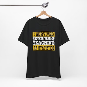 Könnte beinhalten: Schwarzes T-Shirt mit dem Text "I SURVIVED ANOTHER YEAR OF TEACHING AP STATISTICS" in weißer und gelber Schrift. Das Shirt hängt an einem Holzbügel vor weißem Hintergrund.