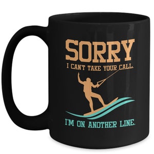 Pode incluir: Caneca de café preta com um design branco. O design diz "SORRY I CAN'T TAKE YOUR CALL. I'M ON ANOTHER LINE." com uma silhueta de uma pessoa praticando wakeboard.