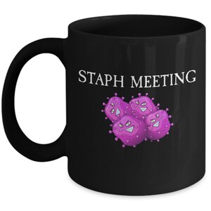 May include: Black coffee mug with a white graphic of purple cartoon bacteria with the text "Staph Meeting" printed on the mug.