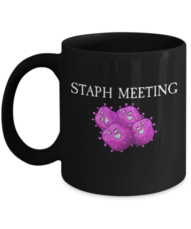May include: Black coffee mug with a white graphic of purple cartoon bacteria with the text "Staph Meeting" printed on the mug.