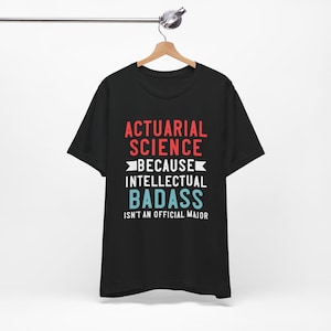 Op de afbeelding: Zwart t-shirt met de tekst "ACTUARIAL SCIENCE BECAUSE INTELLECTUAL BADASS ISN'T AN OFFICIAL MAJOR" in rood, wit en blauw. Het t-shirt hangt aan een houten hanger.