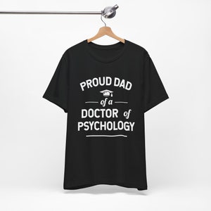 May include: Black t-shirt with white text that reads "Proud Dad of a Doctor of Psychology" with a graduation cap icon.