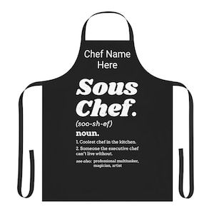 May include: Black apron with white text that reads "Chef Name Here Sous Chef (soo-sh-ef) noun. 1. Coolest chef in the kitchen. 2. Someone the executive chef can't live without. see also: professional multitasker, magician, artist"