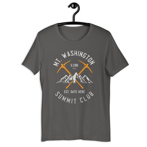May include: A dark grey t-shirt with a white graphic design of a mountain range and two crossed ice axes. The text "MT. WASHINGTON" is above the graphic, and "SUMMIT CLUB" is below. The text "6,288 FEET" is in the centre of the graphic, and "EST. DATE HERE" is below the graphic.