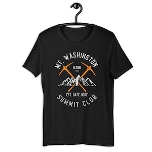 May include: Black t-shirt featuring a white graphic design of a mountain range with two crossed pickaxes. The text "MT. WASHINGTON 6,288 FEET EST. DATE HERE SUMMIT CLUB" is printed on the shirt.