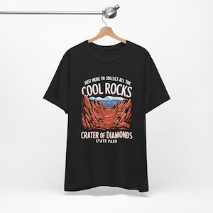 以下が含まれることがあります： 砂漠の風景と「JUST HERE TO COLLECT ALL THE COOL ROCKS CRATER OF DIAMONDS STATE PARK」の文字が描かれた黒いTシャツ。デザインには赤い岩の層、青い空、白い文字が含まれています。