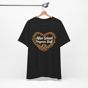 Könnte beinhalten: Schwarzes T-Shirt mit einem herzförmigen Design. Das Design zeigt den Text "After School Program Staff", umgeben von einem Kranz aus Beeren und Blättern, mit einer Kaffeetasse unten.