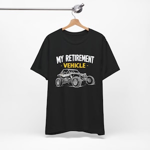 May include: Black t-shirt with the text "MY RETIREMENT VEHICLE" above a white and yellow graphic of an off-road vehicle. The shirt is hanging on a wooden hanger.
