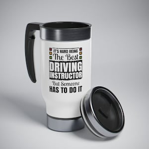May include: White travel mug with a black handle and lid. The mug has a black and white graphic that says "It's hard being the best driving instructor but someone has to do it". There are also three traffic lights in the graphic.