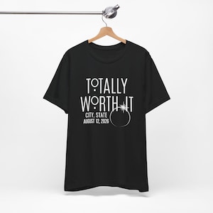 Op de afbeelding: Zwart t-shirt met de witte tekst "TOTALLY WORTH IT", en "CITY, STATE AUGUST 12, 2026". Het t-shirt hangt aan een houten hanger.